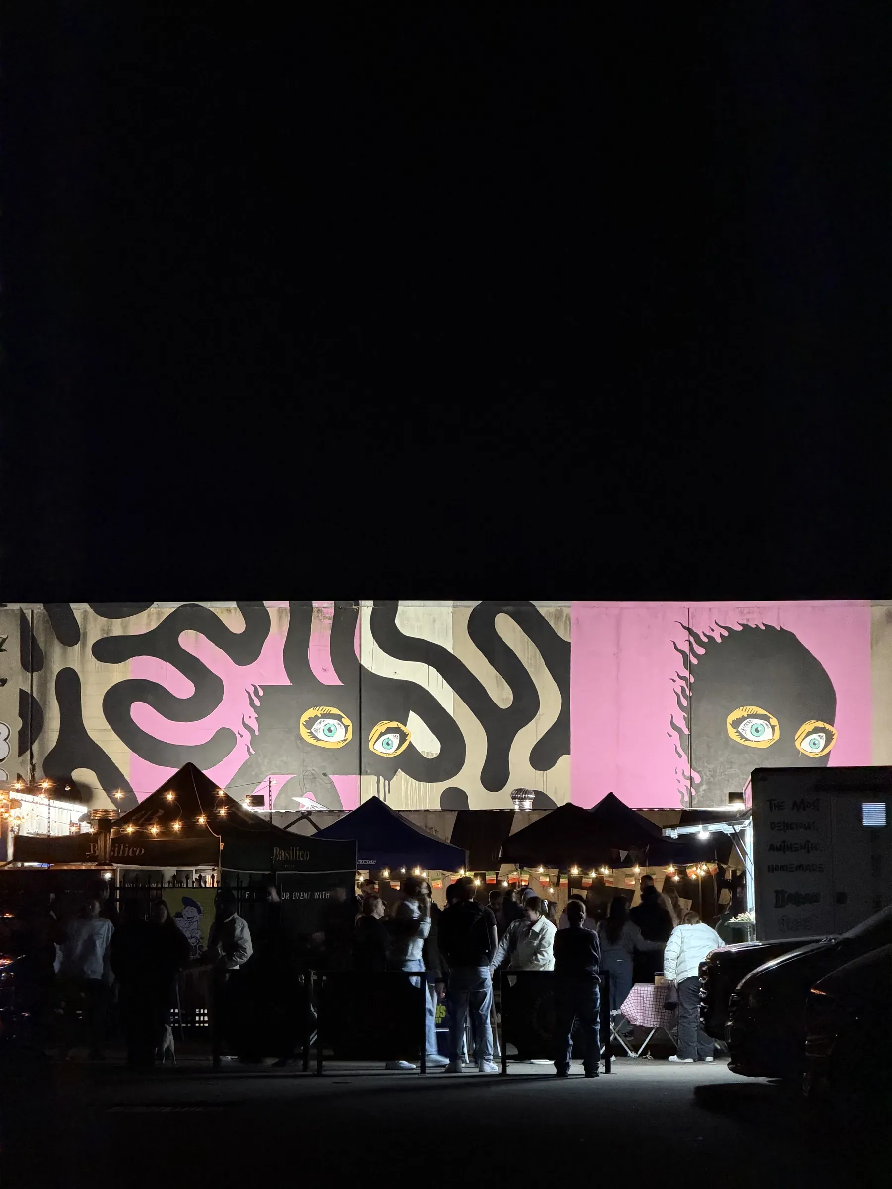 Night view of Toby's Little Italy with Basilico stalls, crowd, and SFB mural under string lights